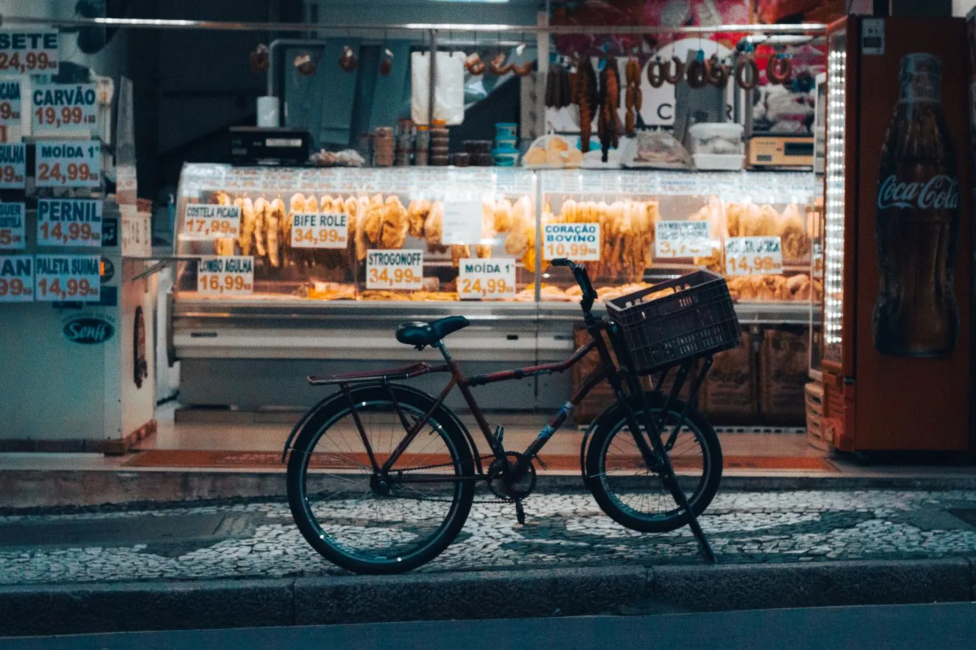 Bicicleta encostada na calçada em frente a um mercado de bairro iluminado, com cartazes de preços de carnes escritos à mão na vitrine.